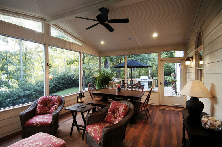 Spacious screened-in porch featuring floral-patterned seating, wooden dining table, and large windows with views of a lush backyard, emphasizing a comfortable outdoor living space.