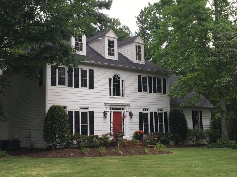 Elegant two-story house with white siding, black shutters, and a red front door, surrounded by lush greenery and landscaped garden, showcasing a classic architectural style ideal for residential settings.