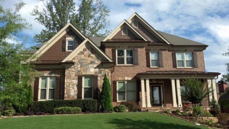 Brick and stone exterior of a two-story home with double-hung windows, landscaped front yard, and blue sky, showcasing the 700 Series vinyl windows from Windowcrafters.