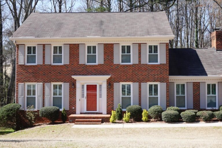 Brick house featuring double-hung windows with white shutters, a central red door, and manicured landscaping, showcasing energy-efficient vinyl windows suitable for residential projects.
