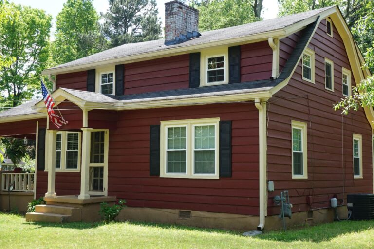 Red two-story house with white double-hung windows, black shutters, and a front porch, surrounded by green grass and trees, showcasing a classic residential design.