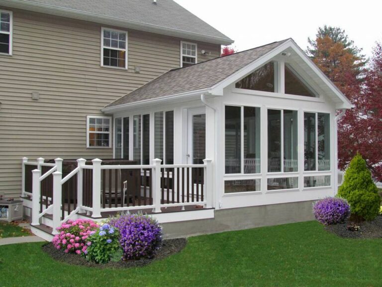 Custom vinyl sunroom addition with large windows, white trim, and a wooden deck, surrounded by colorful flowers and greenery, showcasing Windowcrafters' specialty window solutions for home projects.