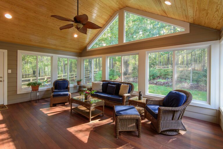 900 Series vinyl windows showcased in a sunlit sunroom with a wooden ceiling, featuring a comfortable seating area, coffee table, and views of a green landscape through large windows.