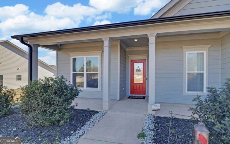 Front entrance of a modern home featuring a red door, flanked by two large windows, with landscaped shrubs and a concrete pathway leading to the entrance.