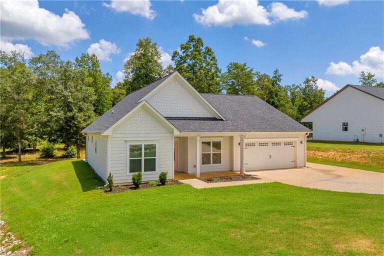 Newly constructed single-family home with a modern design, featuring large vinyl windows, a front porch, and a manicured lawn, surrounded by greenery in a residential neighborhood.