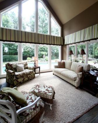 Living room featuring large windows with custom treatments, showcasing natural light and a view of greenery, furnished with a patterned armchair, a sofa, and a decorative ottoman.