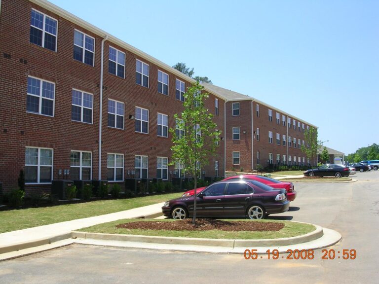 Residential building with multiple stories, featuring brick exterior and several vinyl windows, surrounded by landscaped parking area with parked cars.