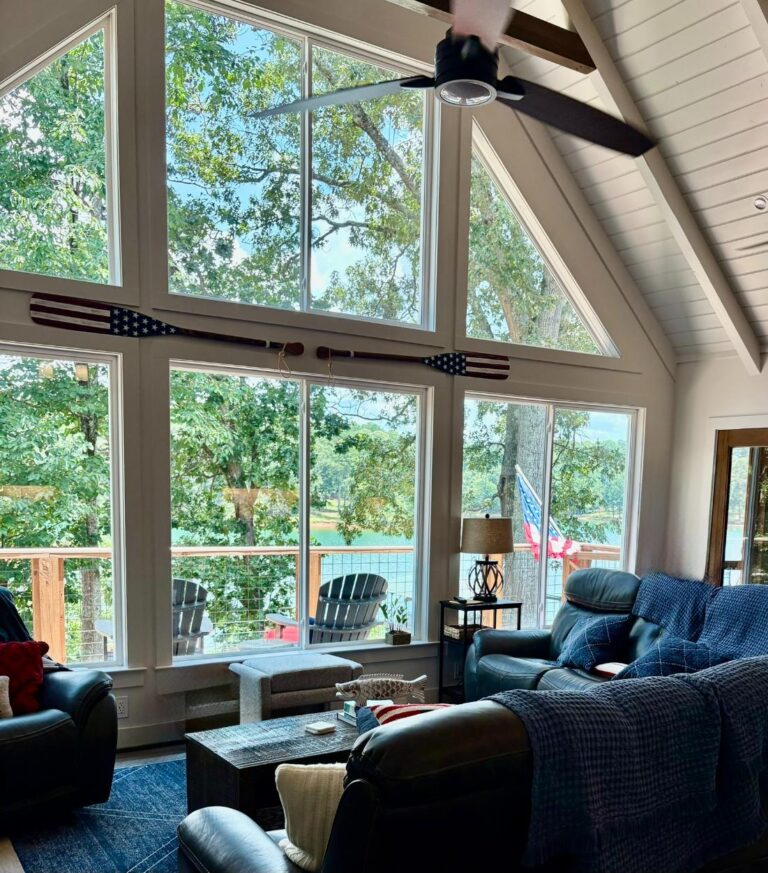 Spacious living room featuring large vinyl windows with a scenic view of trees and water, accented by decorative oars and comfortable seating, highlighting the natural light and open design.