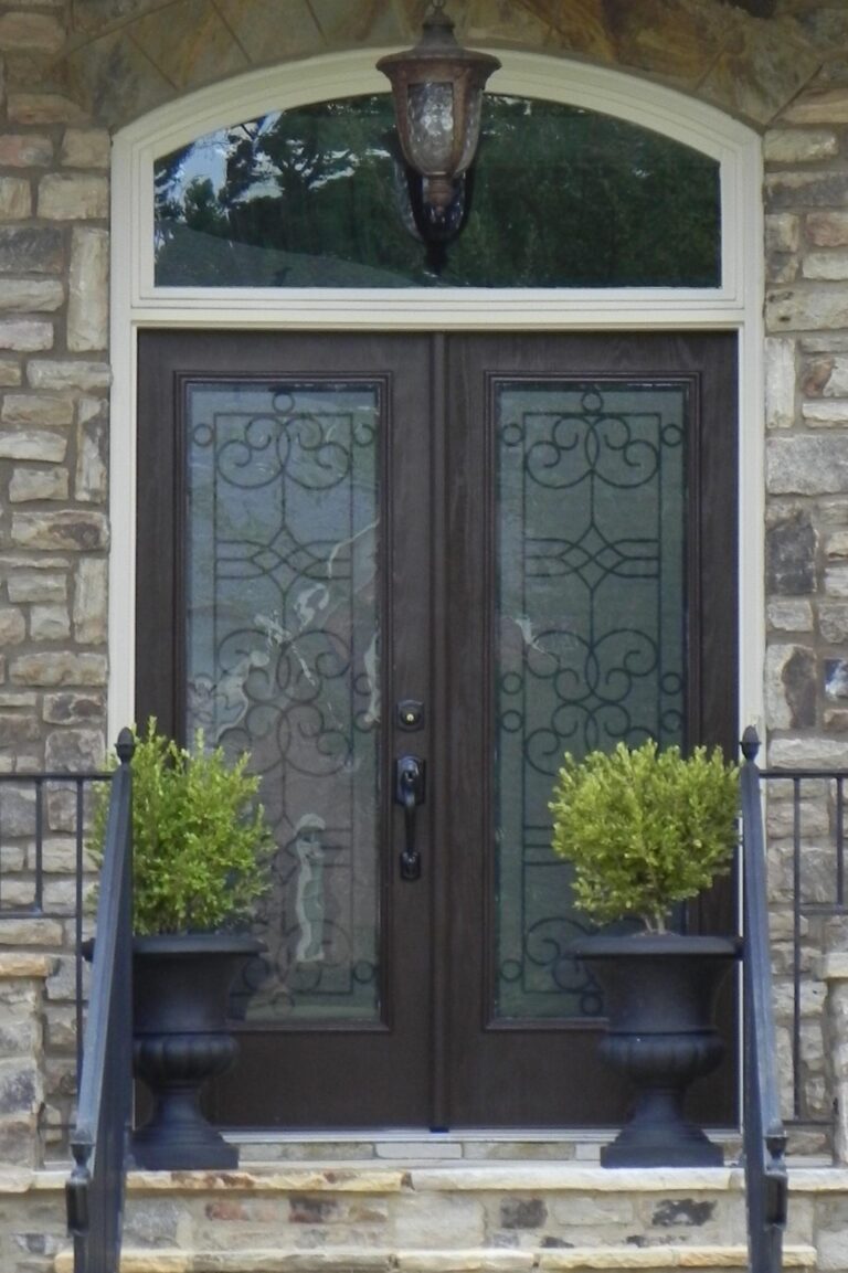 Exterior double doors with decorative glass panels, flanked by potted greenery, set against a stone facade, highlighting quality craftsmanship and design for residential entryways.