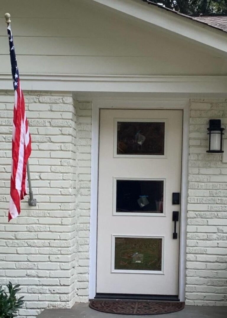Front entrance featuring a modern white exterior door with three glass panels, accompanied by an American flag on a pole and a decorative welcome mat, highlighting options for stylish entryways.