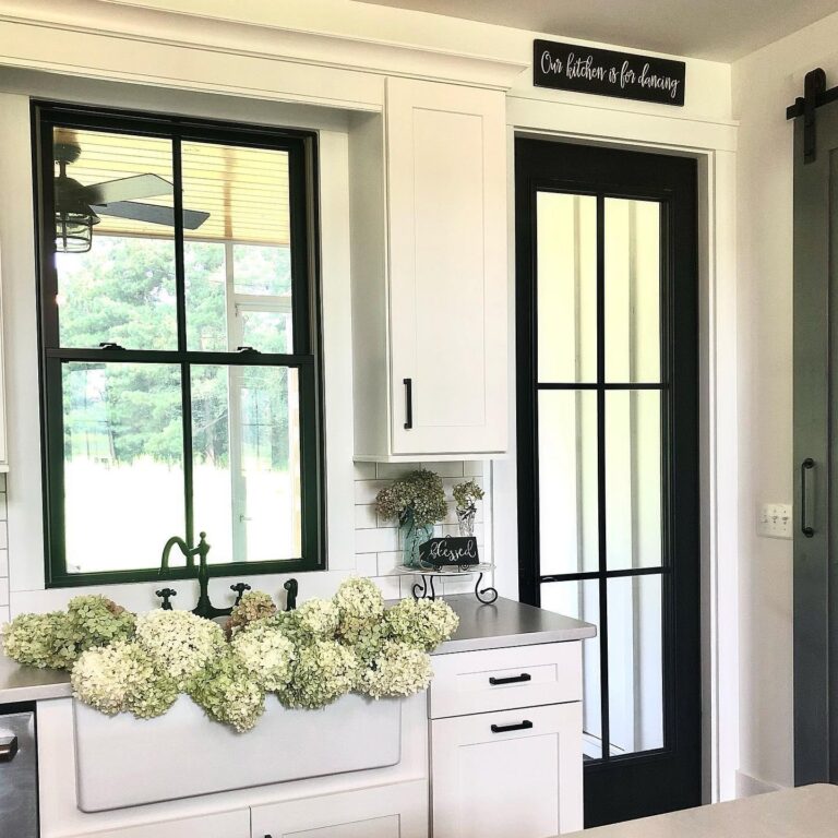Kitchen interior featuring a farmhouse sink adorned with hydrangeas, black-framed windows, white cabinetry, and a black glass door, reflecting a modern yet inviting aesthetic.