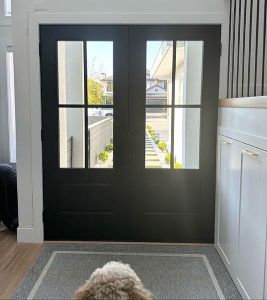 Black double doors with glass panes, leading to a well-lit exterior with a landscaped pathway, showcasing a welcoming entryway for custom and specialty exterior doors.