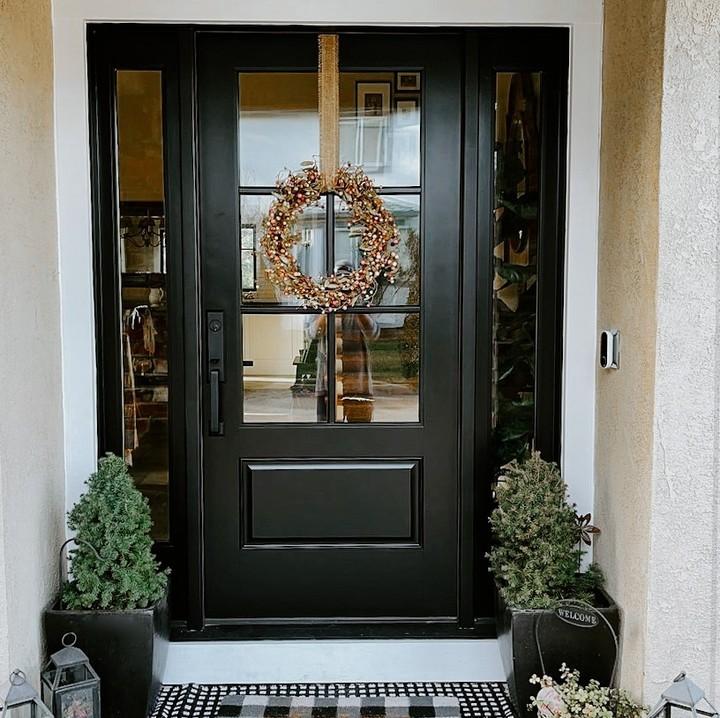 Black entry door with decorative wreath, flanked by potted greenery and a welcome mat, showcasing stylish exterior door design.