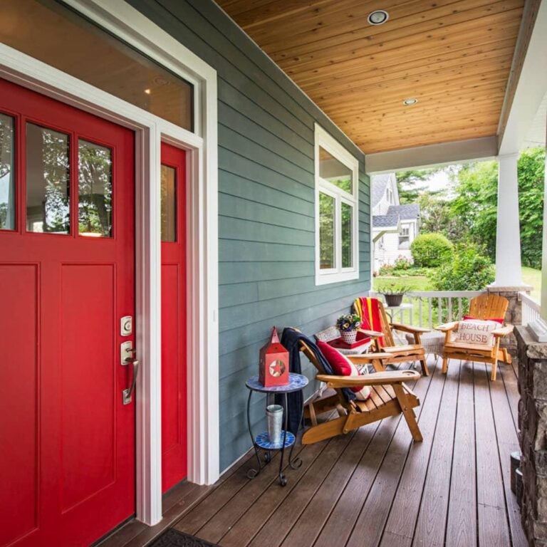 Exterior view of a home showcasing a vibrant red Therma-Tru entry door, surrounded by a welcoming porch with wooden chairs and decorative elements, emphasizing quality and style in exterior door options.