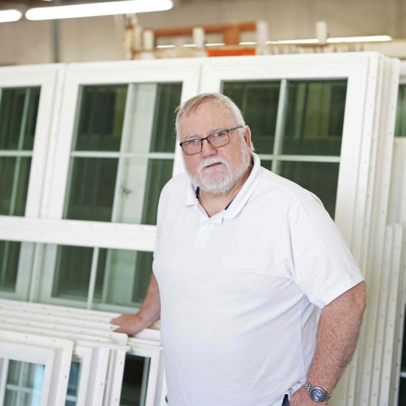 Man in a white polo shirt standing beside vinyl windows in a manufacturing setting, representing Windowcrafters' commitment to quality and local production.