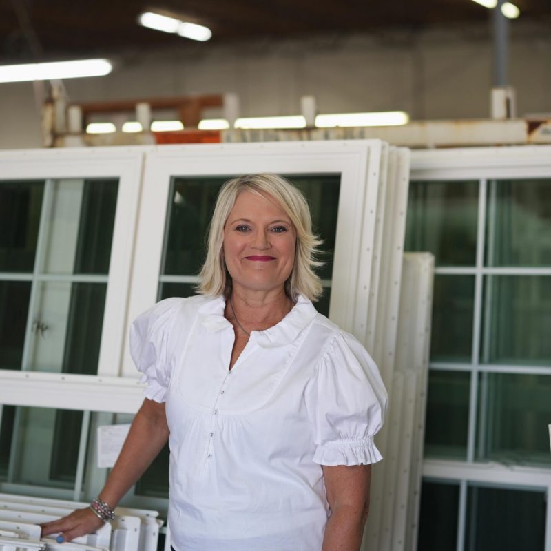 Woman smiling in a factory setting, standing beside vinyl windows, representing Windowcrafters' commitment to quality and professional service in Georgia.
