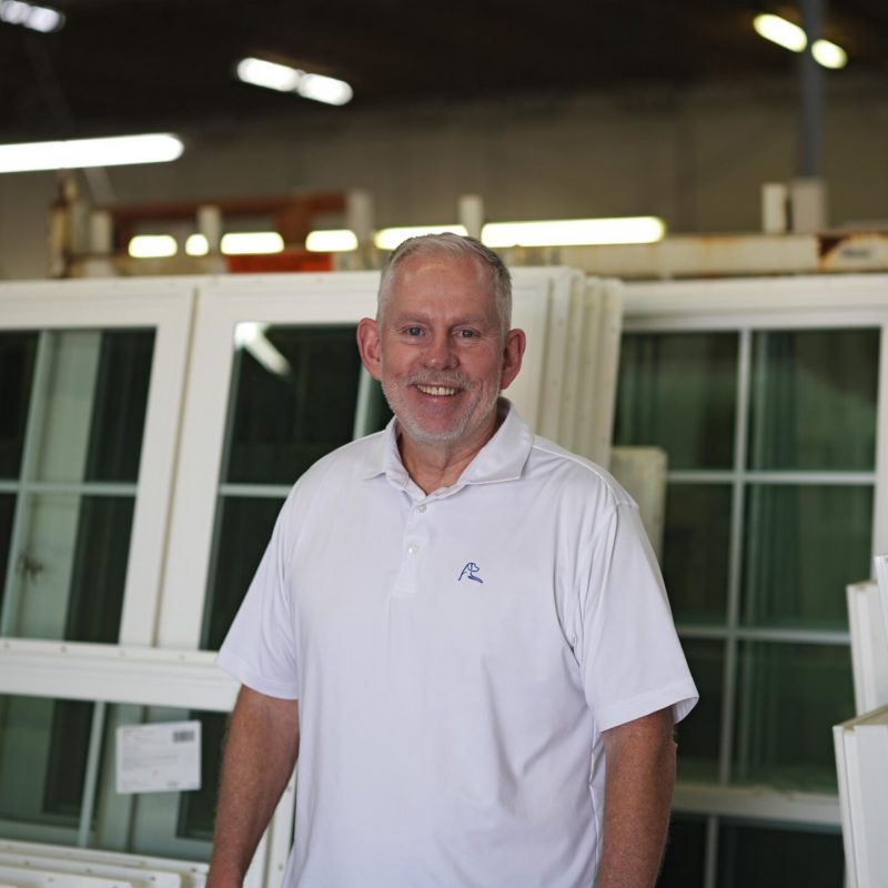 Smiling man in a white polo shirt standing in a factory setting with vinyl windows in the background, representing Windowcrafters' commitment to quality and local manufacturing in Georgia.