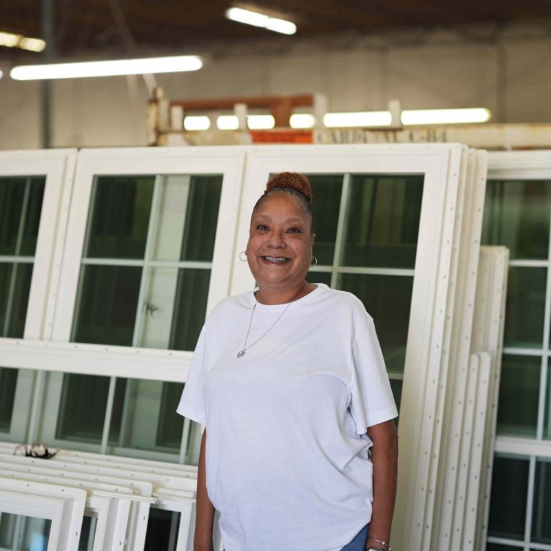 Smiling woman in a white shirt standing in front of vinyl windows in a manufacturing facility, representing Windowcrafters' commitment to quality and local production.