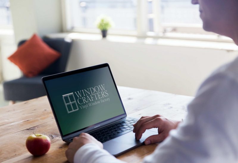 Person using a laptop displaying the Window Crafters logo, with an apple on a wooden table and a cozy interior setting.