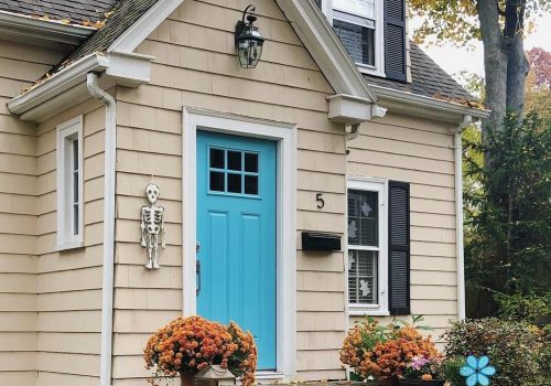Blue entry door with a decorative skeleton, surrounded by colorful flower pots, on a beige house exterior.